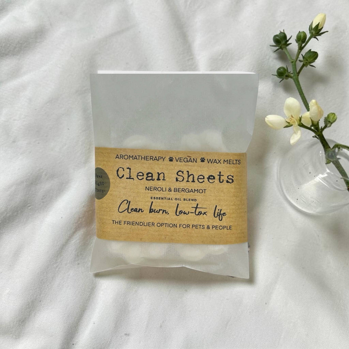 A package of Clean Sheets aromatherapy and vegan wax melts by Meroli & Bergamot, placed on a white fabric background next to a small glass vase with white flowers.