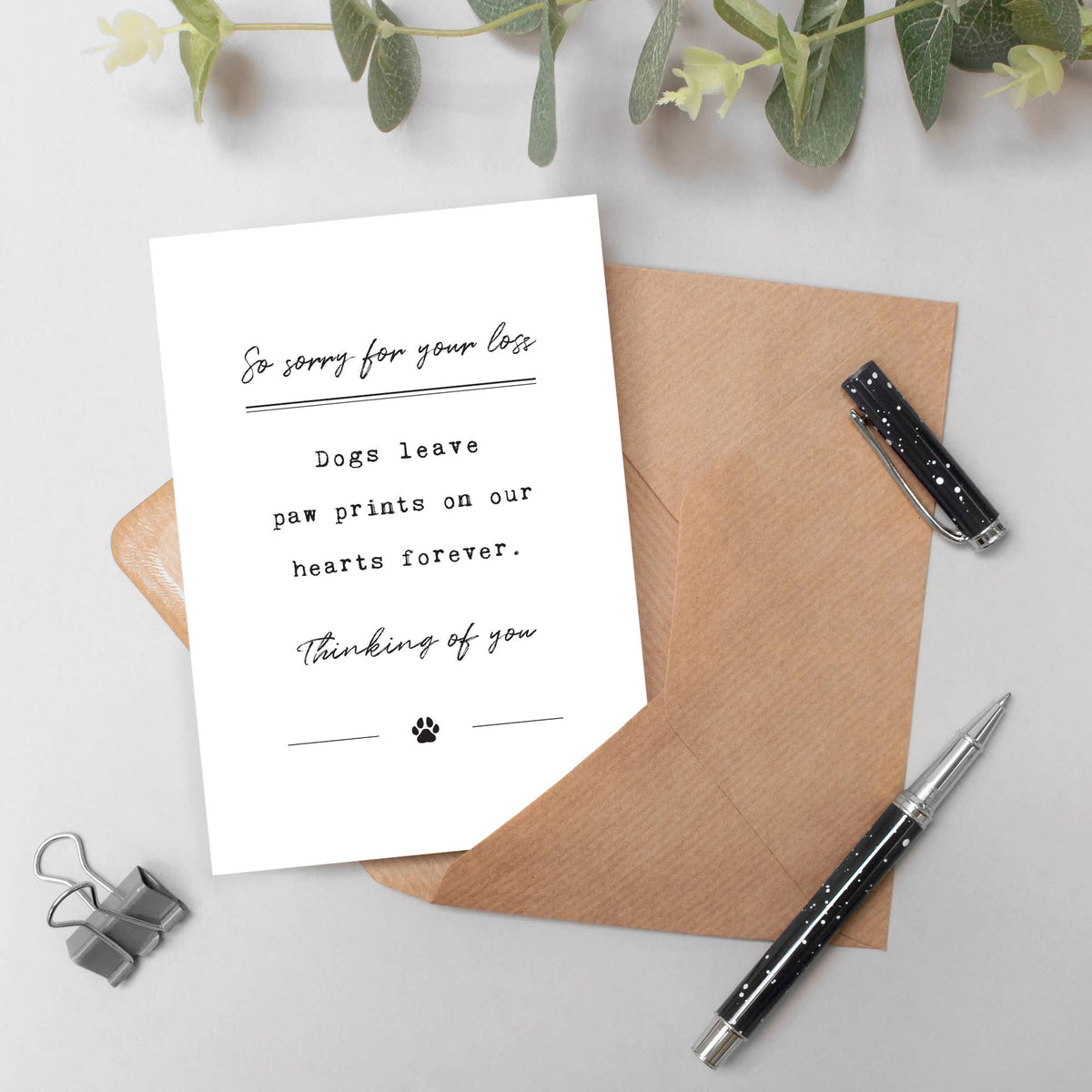 A white greeting card with black script text reading 'So sorry for your loss Dogs leave paw prints on our hearts forever. Thinking of you' with a small paw print, styled on a brown envelope with pens, binder clips, and greenery on a light gray background.